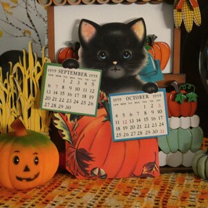 May include: A vintage 1959 calendar featuring a black kitten illustration holding two paper calendar pages for September and October. The display includes orange pumpkin cutouts, a small jack-o'-lantern, and a light green pumpkin figurine.