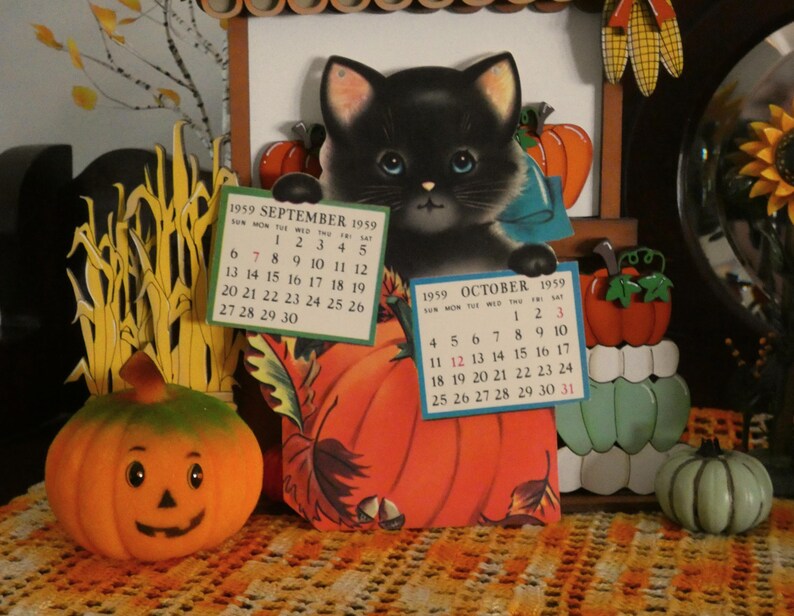 May include: A vintage 1959 calendar featuring a black kitten illustration holding two paper calendar pages for September and October. The display includes orange pumpkin cutouts, a small jack-o'-lantern, and a light green pumpkin figurine.