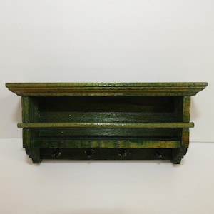 May include: A distressed green wooden wall shelf with a shelf, a rod, and three hooks. The shelf has a decorative edge and is painted in shades of green and brown, giving it a rustic appearance. Ideal for organizing and displaying items.