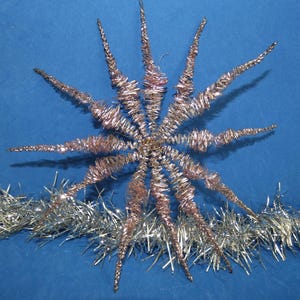 May include: A star-shaped ornament constructed from metallic rose gold wire wrapped in a spiky, textured pattern, featuring twelve radiating points extending from a central core, positioned above a horizontal strand of silver tinsel garland.