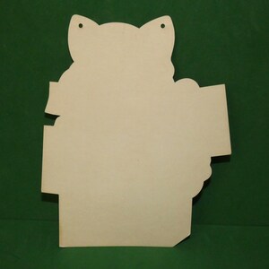 May include: A light beige, die-cut wooden cutout shaped like a cat head with ears and a rectangular sign attached to the side. The surface is smooth and unfinished, featuring two small circular holes at the top for hanging or attachment purposes.