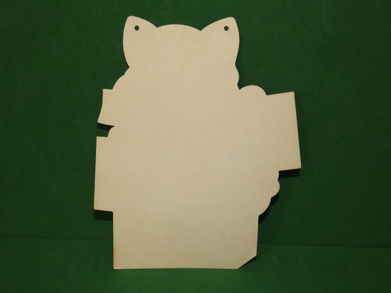 May include: A light beige, die-cut wooden cutout shaped like a cat head with ears and a rectangular sign attached to the side. The surface is smooth and unfinished, featuring two small circular holes at the top for hanging or attachment purposes.