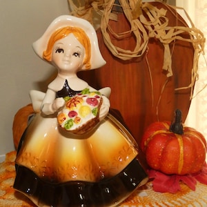 May include: A ceramic figurine of a person with orange hair wearing a white bonnet and a dress that fades from white to orange and dark brown at the hem. The figure holds a basket filled with colorful painted fruits and vegetables in their hands.