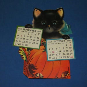 May include: A vintage 1959 paper calendar featuring a black kitten with blue eyes holding two rectangular calendar pages for September and October. The kitten sits behind a large orange pumpkin decorated with autumn leaves and small brown acorns.