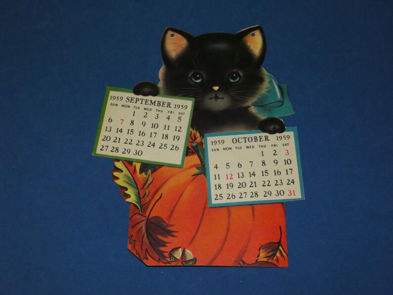 May include: A vintage 1959 paper calendar featuring a black kitten with blue eyes holding two rectangular calendar pages for September and October. The kitten sits behind a large orange pumpkin decorated with autumn leaves and small brown acorns.