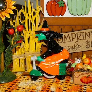 May include: A decorative witch figurine with green skin and a black hat riding a green scooter, positioned next to a sunflower, a wooden corn stalk cutout, a sign reading PUMPKINS sale $5, and a small crate labeled HANDPICKED PUMPKINS.
