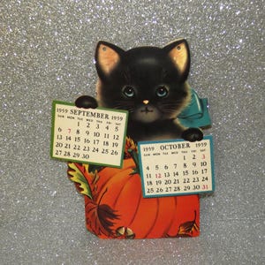 May include: A vintage 1959 paper calendar featuring a black kitten with blue eyes holding two rectangular cards. The left card displays September 1959 and the right card displays October 1959. The kitten sits behind a large orange pumpkin with leaves.