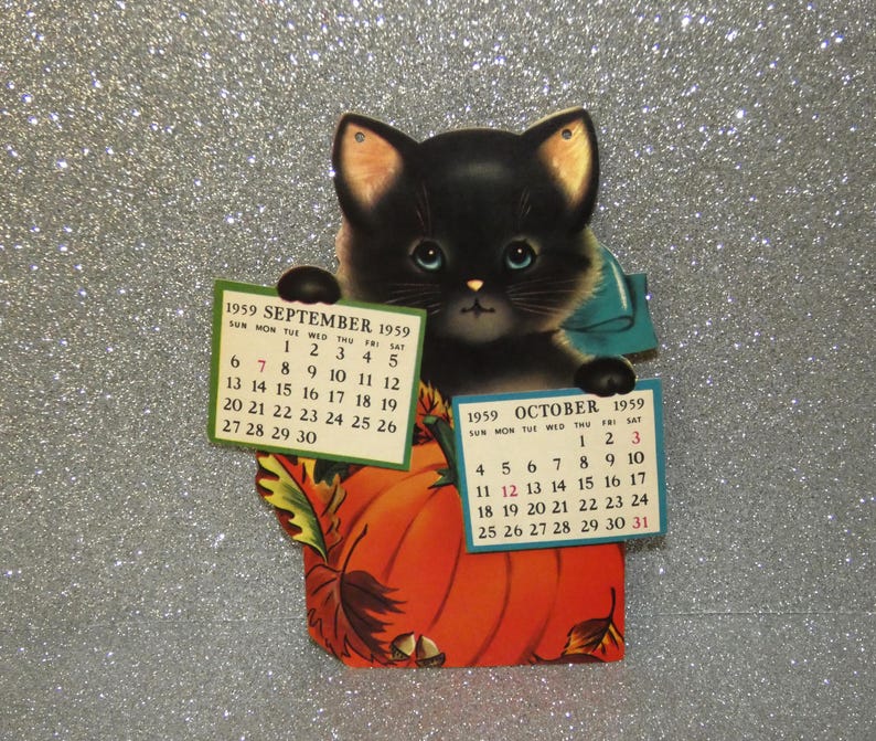 May include: A vintage 1959 paper calendar featuring a black kitten with blue eyes holding two rectangular cards. The left card displays September 1959 and the right card displays October 1959. The kitten sits behind a large orange pumpkin with leaves.