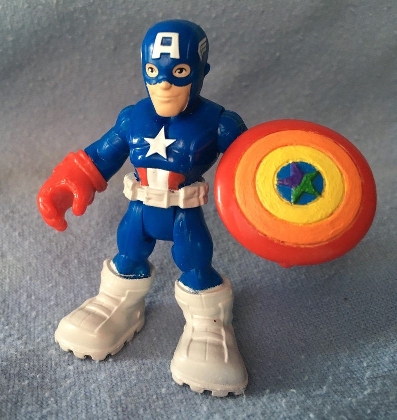 May include: A Captain America action figure in a dynamic pose, clad in a blue suit with a white star, red gloves, and white boots. The figure holds a colorful shield with a star emblem, a classic superhero toy.