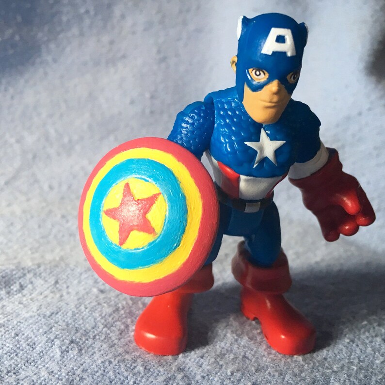 May include: A Captain America action figure. The figure is wearing a blue helmet with a white "A", a blue suit with a white star, red gloves and boots. It holds a shield with red, yellow, and blue concentric circles and a red star.