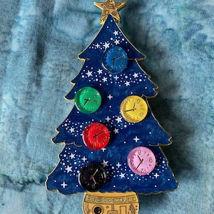 May include: A whimsical Christmas tree-shaped pin with a dark blue background, adorned with colorful clock faces. The tree features a gold star and base, with white star accents. The clock faces are in various colors: blue, green, red, black, yellow, and pink.