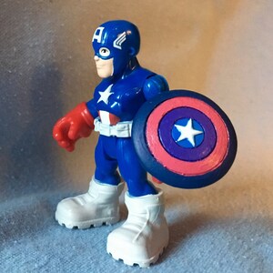 May include: A blue Captain America action figure with a white star on the chest and shield. The figure has a blue helmet with a white "A" and wings, red gloves, and white boots. The shield is red, white, and blue.