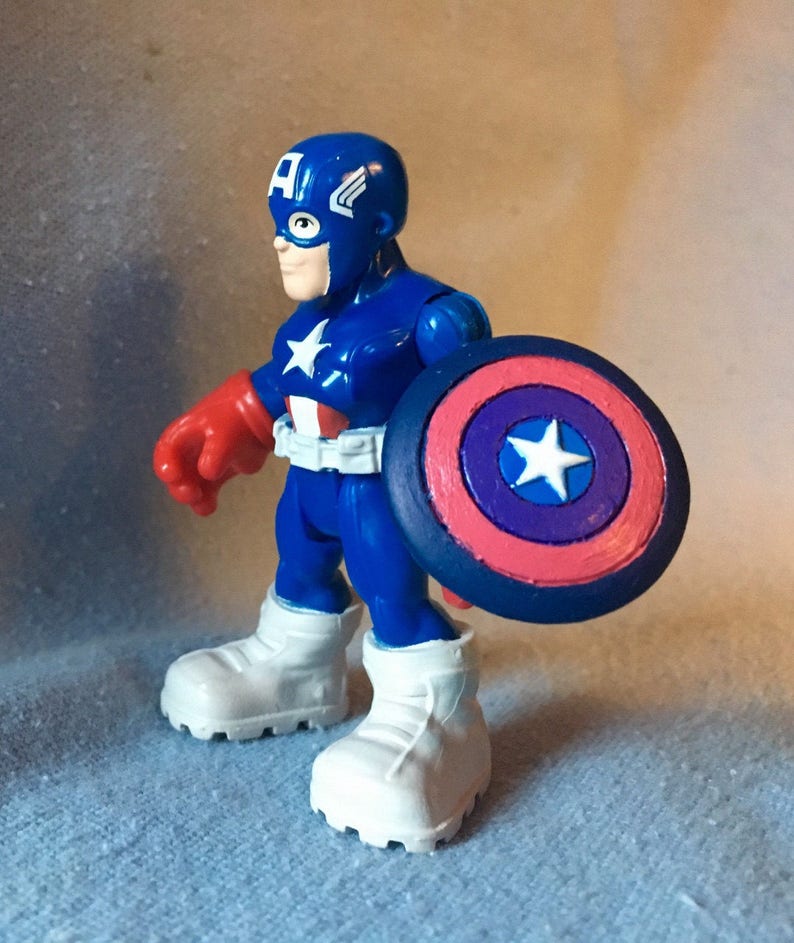 May include: A blue Captain America action figure with a white star on the chest and shield. The figure has a blue helmet with a white "A" and wings, red gloves, and white boots. The shield is red, white, and blue.