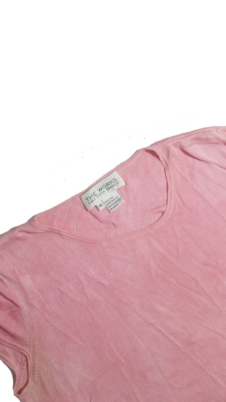 May include: A pink, short-sleeved, ribbed knit top with a white tag that reads "THE WORKS SAKS FIFTH AVENUE" and "100% Silk Made in China Dry Clean Only M (US) 00237"