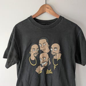 May include: Black t-shirt featuring a cartoon graphic of four men with gold jewelry and the text "fresh talent". The design is in shades of brown, tan, and gold. The shirt is hanging on a wooden hanger.