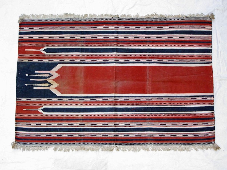 Puede incluir: Una alfombra de rayas rojas, blancas y azules con un panel central rojo. La alfombra tiene un borde de flecos y est&aacute; tejida con patrones intrincados.