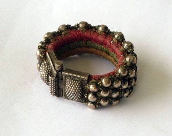 Puede incluir: Un brazalete de plata con una banda de tela roja y verde. El brazalete está decorado con pequeñas cuentas de plata y tiene un cierre.