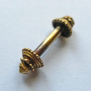 May include: A small, ornate, gold-colored metal object with a cylindrical body and decorative, circular ends. The ends feature intricate beaded detailing and a pointed tip on one end. The object is likely a decorative element or jewelry component.