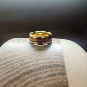 May include: A gold and brown ring with a wood inlay design. The ring is sitting on a wooden surface.