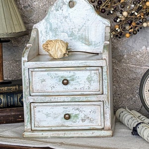 May include: A small, white, two-drawer cabinet with a distressed finish. The cabinet has a decorative top with a scalloped edge and a small hole in the centre. The cabinet is sitting on a table with a white tablecloth.