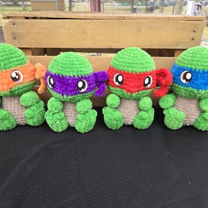 May include: Four crocheted Teenage Mutant Ninja Turtles, each with a different colored mask: orange, purple, red, and blue. The turtles are sitting on a black surface.