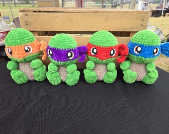 May include: Four crocheted Teenage Mutant Ninja Turtles, each with a different colored mask: orange, purple, red, and blue. The turtles are sitting on a black surface.