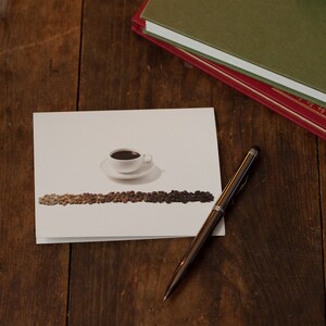 May include: A white card with a cup of coffee and a line of coffee beans printed on it. A silver pen with a black stylus tip is lying on a wooden surface next to the card.