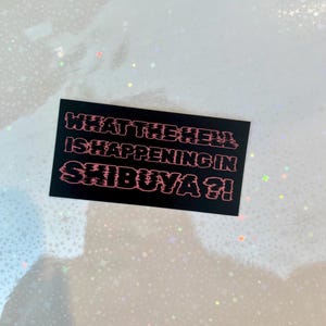 May include: A black rectangular sticker with the text "WHAT THE HELL IS HAPPENING IN SHIBUYA?!" in pink, glitch-style font. The sticker is set against a background with a sparkly, iridescent effect.