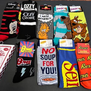 May include: A selection of crew socks with diverse designs. The socks feature characters like Mickey Mouse, Ozzy Osbourne, Garfield, Scooby-Doo, David Bowie, and Seinfeld. Also includes Reese's peanut butter cup socks.
