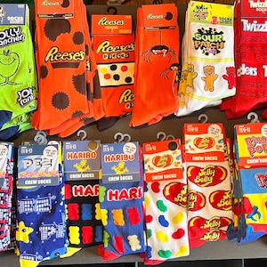 May include: A collection of colourful crew socks featuring various candy brands, including Jolly Rancher, Reese's, Sour Patch Kids, Twizzlers, Ring Pop, and Jelly Belly. The socks are displayed on a black surface.
