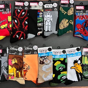 Puede incluir: Una colección de 16 calcetines de tripulación coloridos con varios personajes de dibujos animados y logotipos. Los calcetines se muestran en una superficie blanca. Los personajes incluyen Deadpool, Ironman, Groot, Spiderman, Homer Simpson, Mickey Mouse, Simba, Bugs Bunny y Hulk.