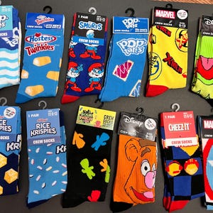 May include: A collection of 10 pairs of crew socks featuring various popular brands and characters. The socks are displayed on a white surface. The brands include Pop-Tarts, Twinkies, Smurfs, Rice Krispies, Sour Patch Kids, Cheez-It, and Marvel. The characters include Smurfs, a green monster, and a cartoon bear.