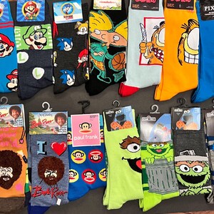 May include: A collection of colorful crew socks featuring various cartoon characters, including Mario, Sonic, Garfield, and Bob Ross. The socks are displayed on a white surface.
