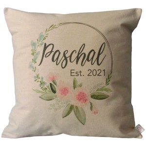 Op de afbeelding: Een beige kussen met een bloemenkransontwerp. De krans is in tinten groen, roze en blauw. De tekst "Paschal Est. 2021" is in zwart binnen de krans gedrukt.