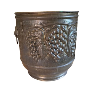 Peut inclure: Un seau cylindrique en métal avec une patine sombre et vieillie, présentant un relief embossé proéminent de grappes de raisin et de feuilles de vigne sur tout l'extérieur. Le récipient comprend un bord robuste et une attache de poignée latérale.