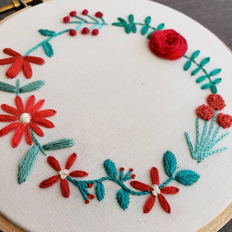 以下が含まれることがあります： 赤い糸とターコイズブルーの糸で手縫いされた刺繍枠。花輪のデザインで、花、葉、ベリーが描かれています。