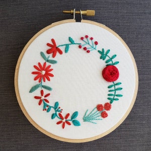 以下が含まれることがあります： 赤と緑の花柄の白い刺繍枠。デザインは、赤い花と緑の葉が円形に配置されています。