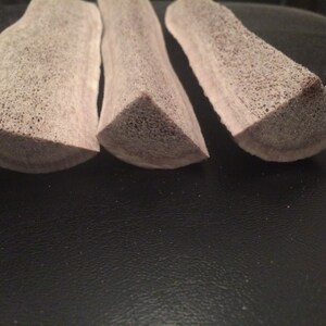 May include: Three light brown antler chews, each with a triangular cross-section, are arranged in a row on a dark surface.