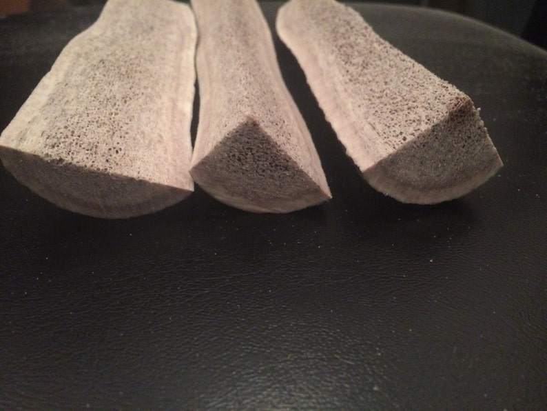 May include: Three light brown antler chews, each with a triangular cross-section, are arranged in a row on a dark surface.