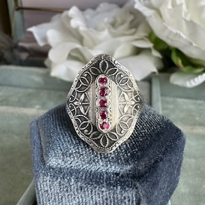 May include: A silver ring with a filigree design and a row of pink gemstones set in the center.