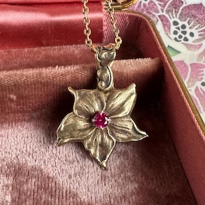 May include: A gold-toned pendant necklace featuring a flower design with a central red gemstone. The flower has detailed petals and is suspended from a delicate gold chain. The necklace is displayed inside a pink jewelry box.