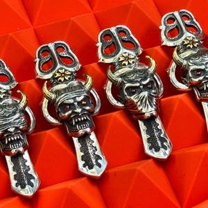 May include: Four silver pendants with a demonic face design, featuring horns, red eyes, and intricate detailing. The pendants have a stylized top and are set against a red geometric background.