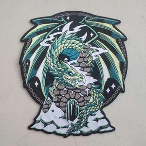 May include: Embroidered patch featuring a green and teal dragon coiled around a stone structure. The dragon has detailed scales and a fierce expression. The patch has a black border with a chain design and star accents, set against a white background.