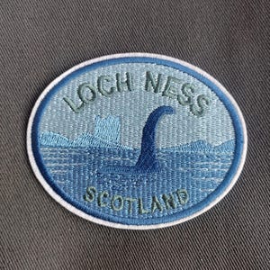May include: Embroidered patch featuring the Loch Ness Monster in shades of blue, with the words "LOCH NESS SCOTLAND" in green lettering. The patch is oval-shaped with a white border, set against a gray background.