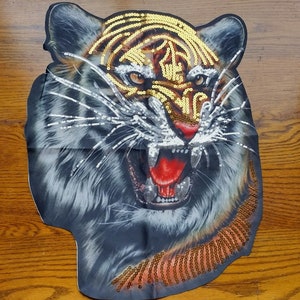 May include: A close-up of a tiger's face with gold and silver sequins. The tiger is roaring with its mouth open and teeth showing. The background is a dark wood grain.
