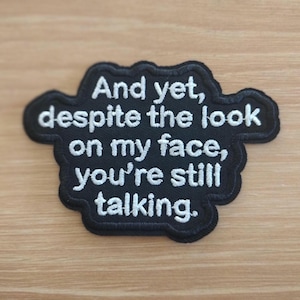 May include: Black embroidered patch with white text that reads "And yet, despite the look on my face, you're still talking." The patch has a wavy border and is displayed on a light brown surface.