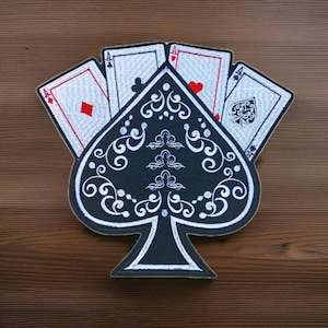 May include: An embroidered patch featuring a large black spade symbol decorated with white filigree patterns, layered over four white playing cards. Each card displays an ace with its respective suit symbol in red or black, including diamonds, clubs, hearts, and spades.