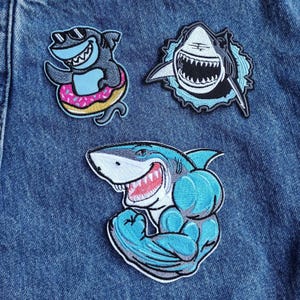 May include: Three embroidered iron-on patches featuring sharks. One patch shows a shark wearing sunglasses and holding a pink donut. Another patch shows a white shark with a wide open mouth. The third patch shows a blue shark with a muscular arm.