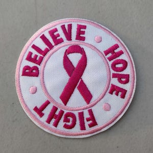 May include: Round white embroidered patch with a pink ribbon in the center. The words "BELIEVE", "HOPE", and "FIGHT" are embroidered in pink around the ribbon. The patch has a pink border and small pink dots.