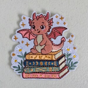 May include: An embroidered patch featuring a small pink dragon with wings sitting atop a stack of four colorful books. The design includes white daisy flowers on the sides and gold embroidered stars scattered across the light blue background.